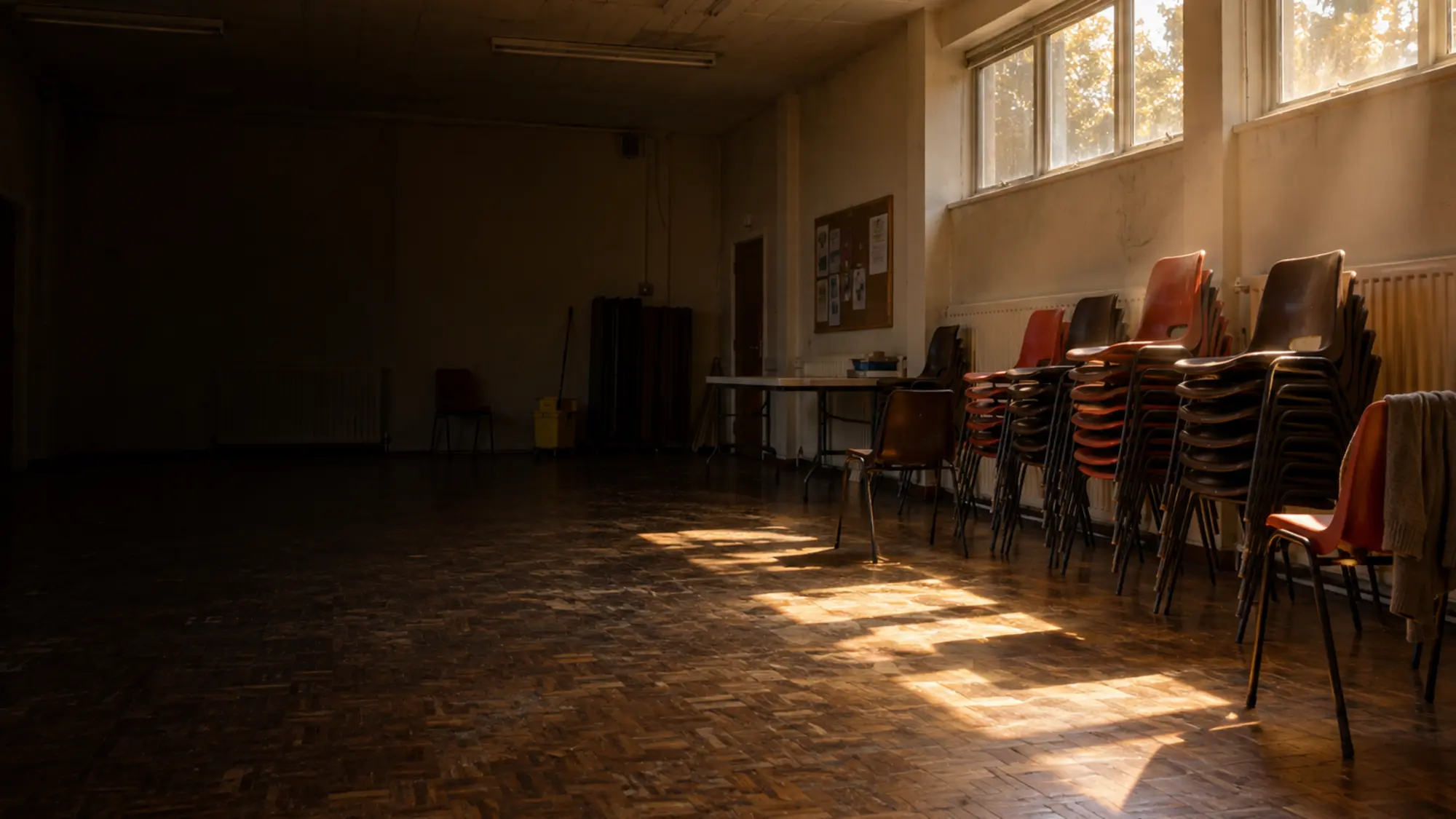 An empty community hall with stacked chairs, late afternoon light coming through the windows